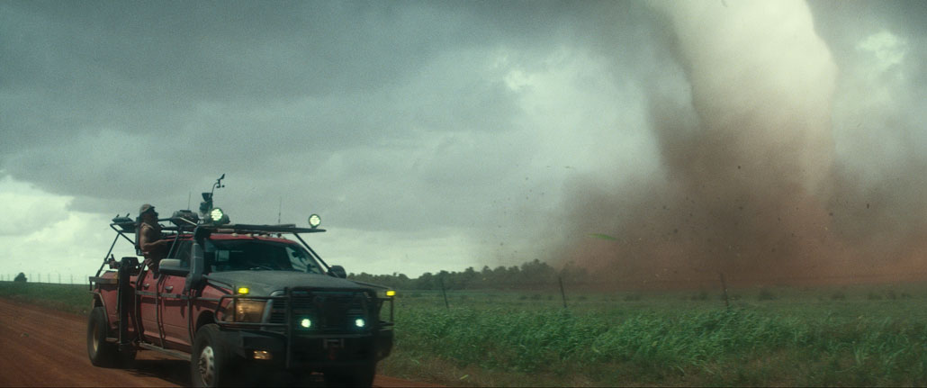 A vertical shot from the movie 'Twisters' shows a red truck on the right equipped with tornado tracking and additional lights.  A man leans halfway out the window to look toward a nearby tornado on the right side of the image.